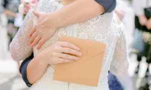 Dłoń trzymająca kopertę na tle sukni ślubnej i uścisku.